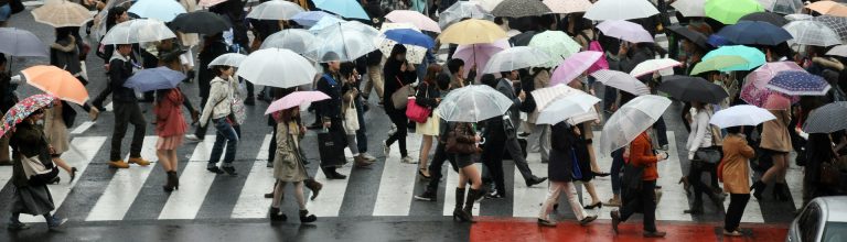 Crowded city crosswalk in heavy rain with hundreds of pedestrians holding umbrellas, traffic waiting, and a bus approaching on wet street.