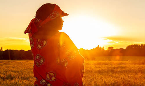 African woman in traditional dress shading eyes in wheat field at sunset panorama.
