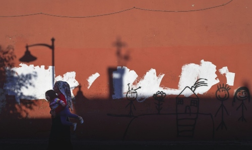 A woman carrying a young child walks past an orange wall with white patches and simple stick-figure graffiti, including drawings of people, a flower, and a house